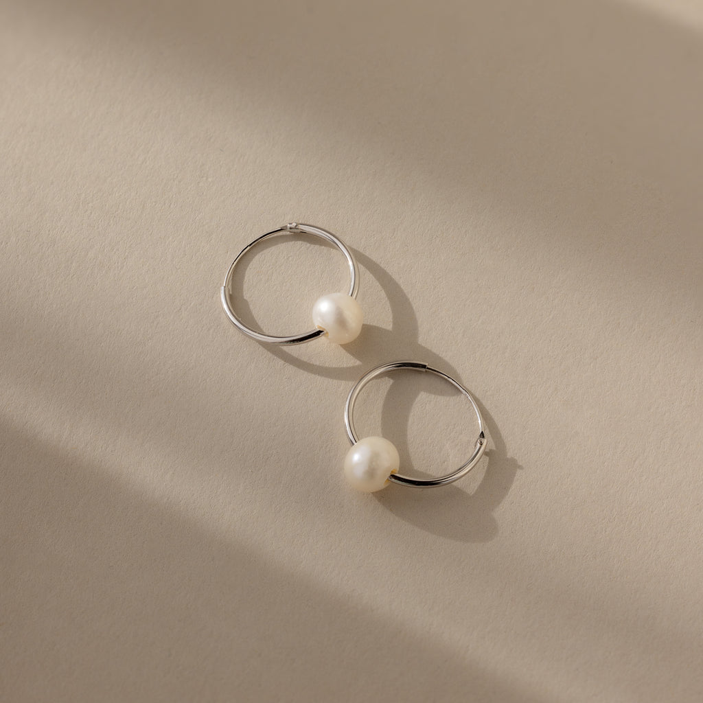 The Round Pearl Hoops in Sterling Silver, showcasing polished sterling silver hoops each adorned with a luminous pearl, are displayed on a beige surface with soft shadows.