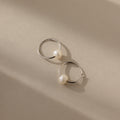 The Round Pearl Hoops in Sterling Silver, showcasing polished sterling silver hoops each adorned with a luminous pearl, are displayed on a beige surface with soft shadows.