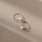 The Round Pearl Hoops in Sterling Silver, showcasing polished sterling silver hoops each adorned with a luminous pearl, are displayed on a beige surface with soft shadows.