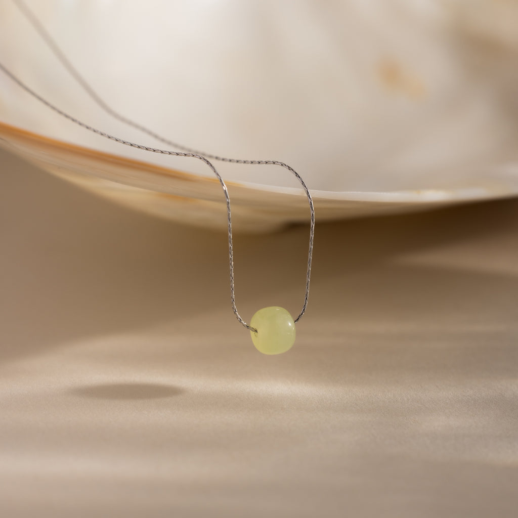 Close-up of a delicate silver chain necklace featuring a single round green jade bead, draped from a white shell dish.
