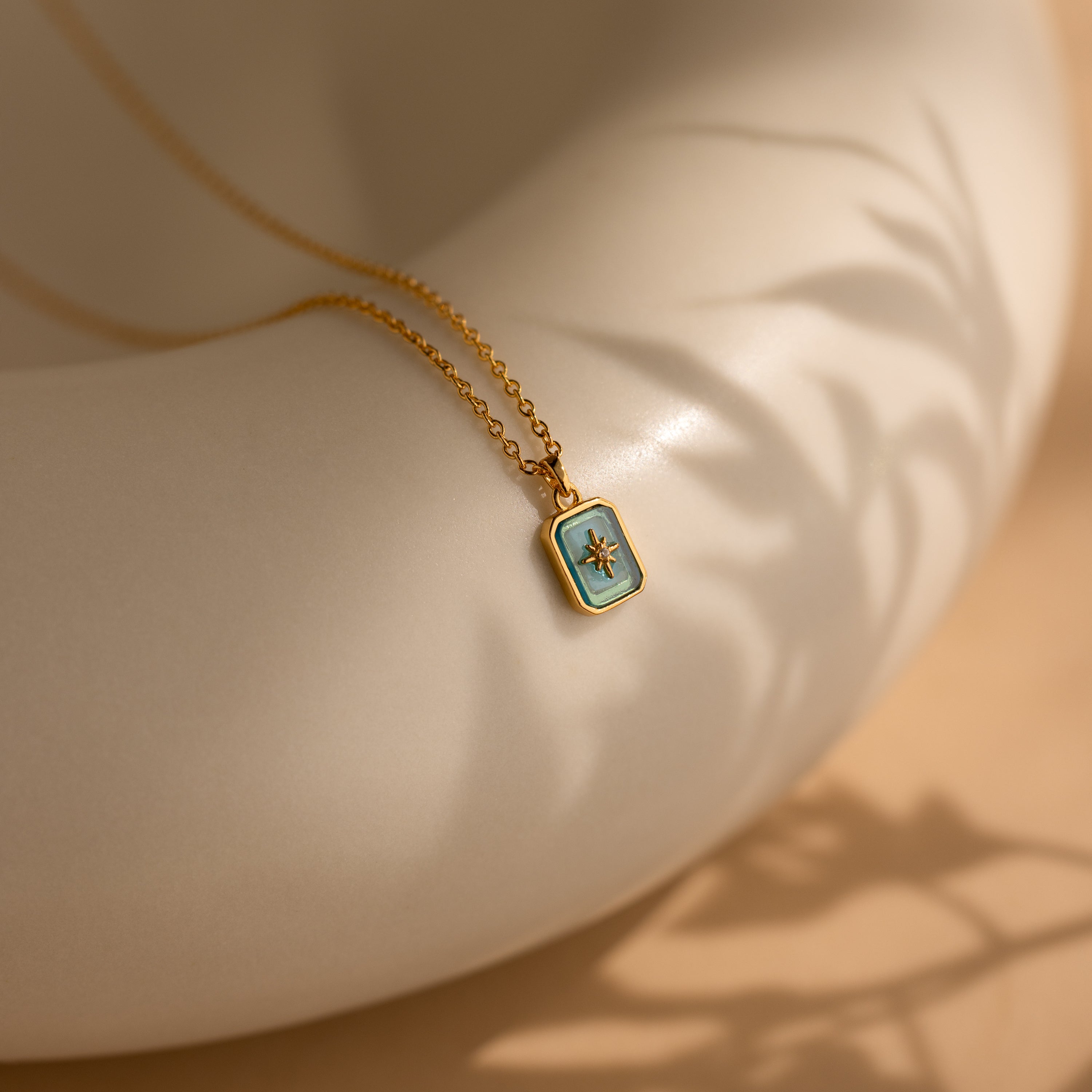 The North Star Birthstone Necklace in blue zircon (December) features a rectangular pendant resting on a curved cream surface with delicate leaf shadows.