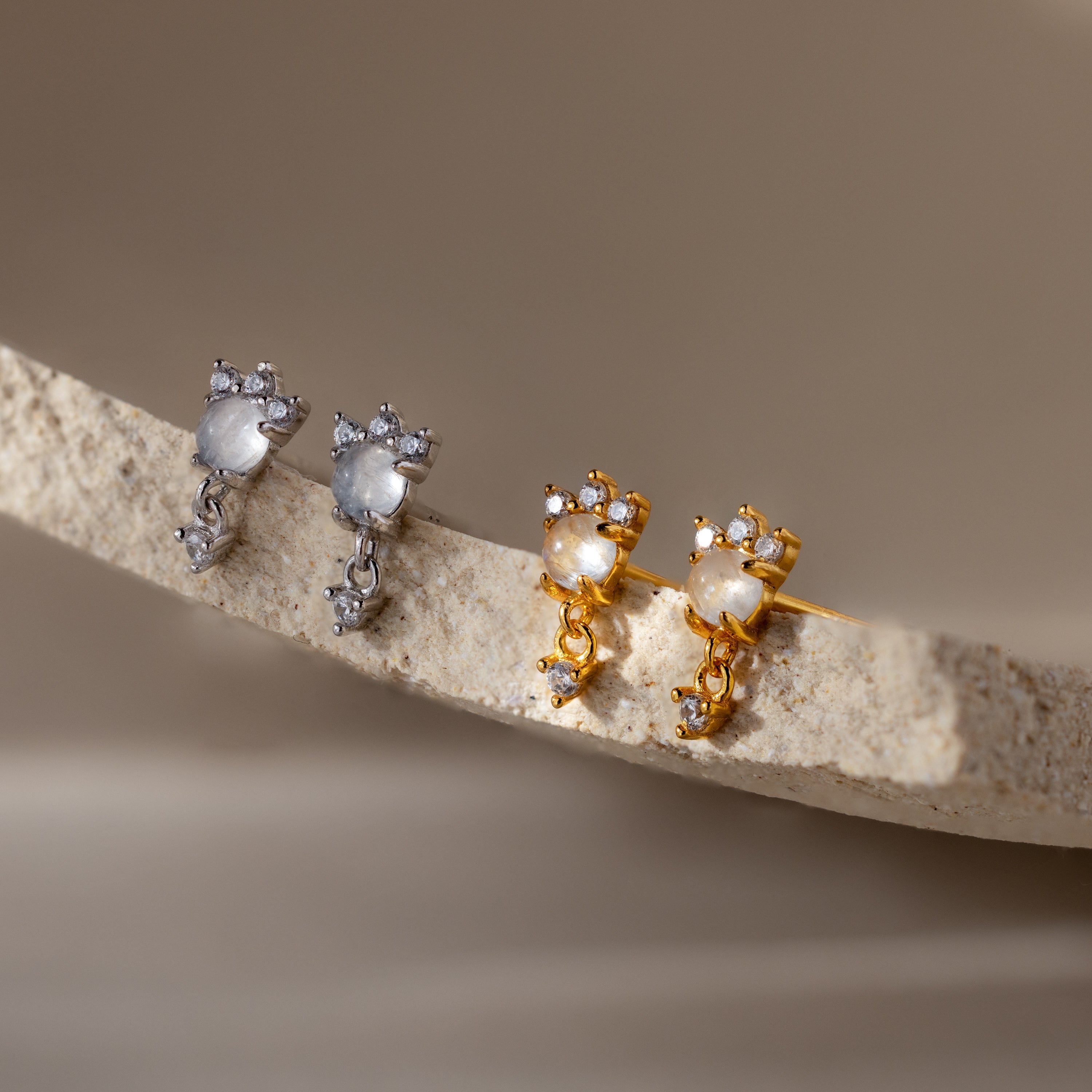 Two pairs of Halo Moonstone Drop Studs, one silver and one gold, displayed on a textured beige surface—perfect for mixing with your favorite diamond studs.