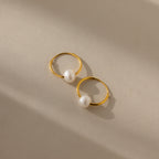 The Round Pearl Hoops in 18K Gold, featuring single white pearls, rest on a beige surface under soft light and shadow, embodying timeless minimal jewelry elegance.