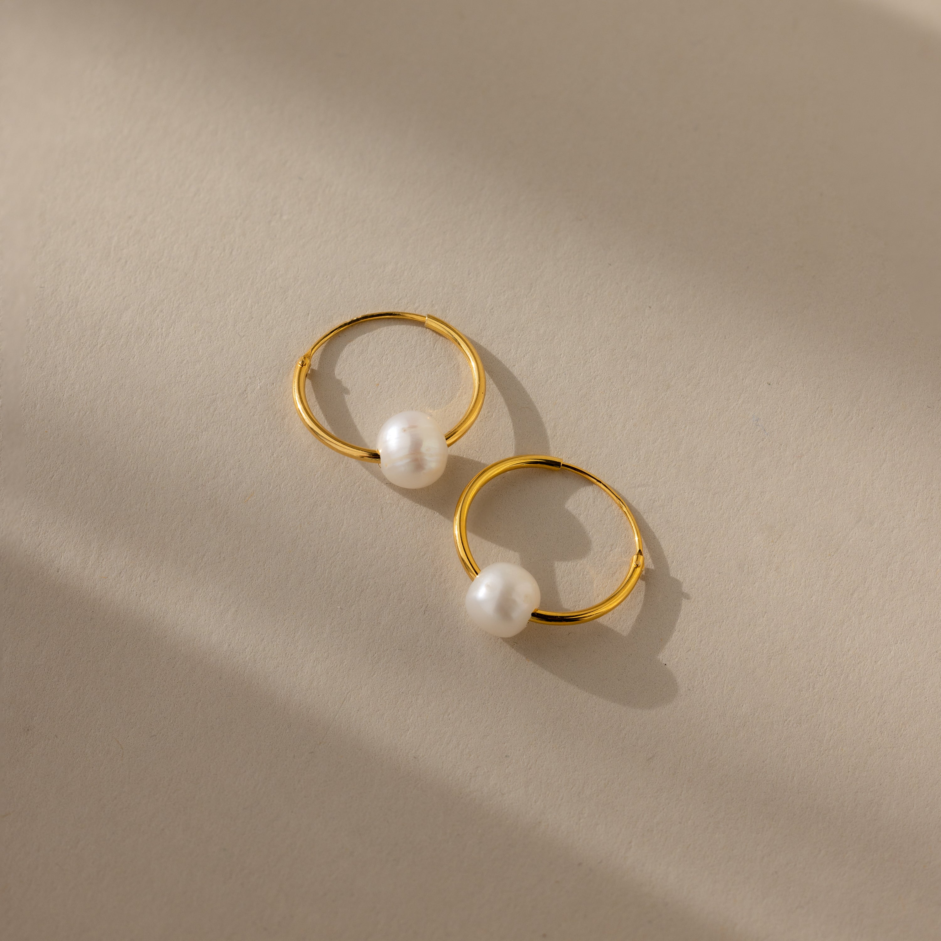 The Round Pearl Hoops in 18K Gold, featuring single white pearls, rest on a beige surface under soft light and shadow, embodying timeless minimal jewelry elegance.