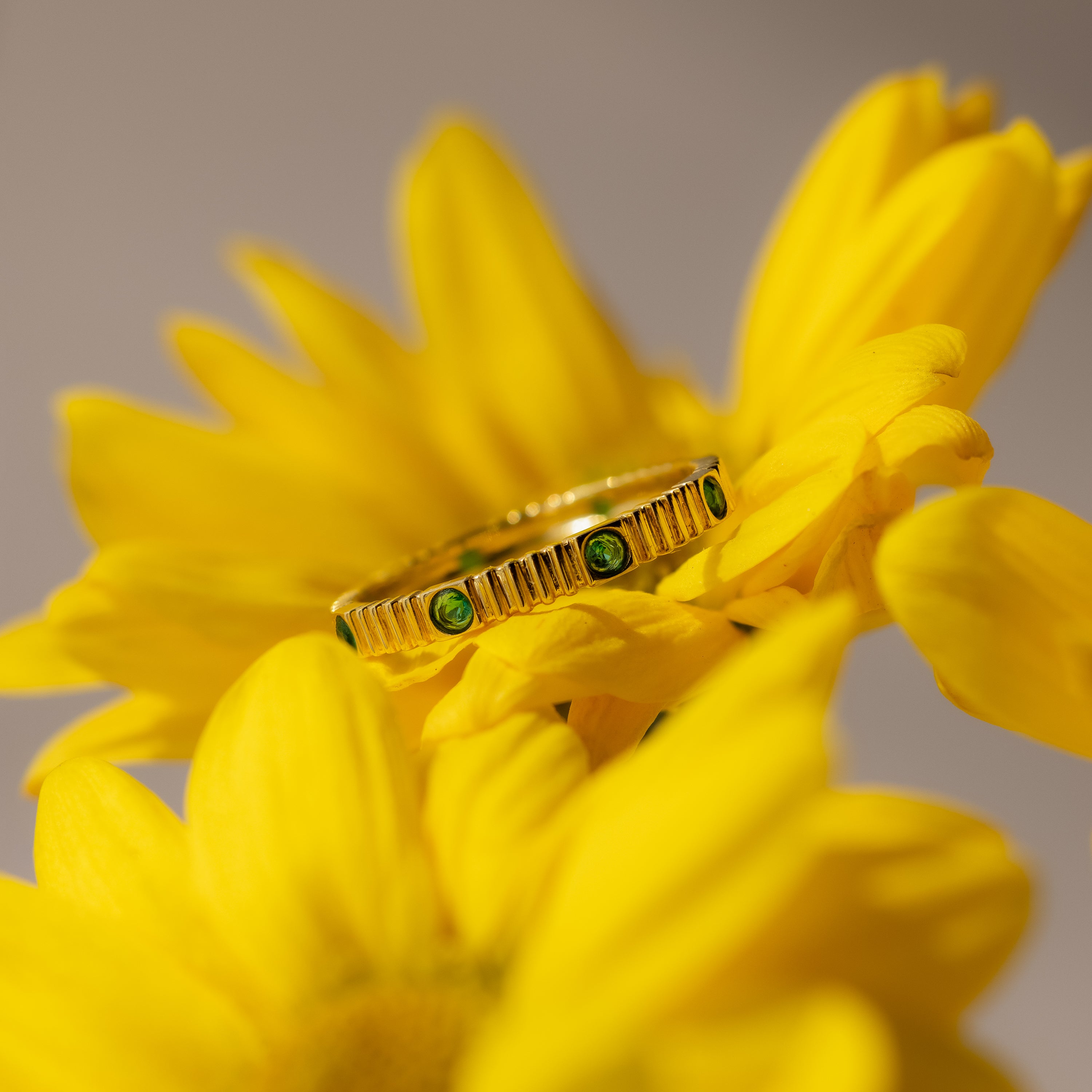 The Ribbed Round Birthstone Eternity Ring with green gemstones rests on bright yellow flower petals.