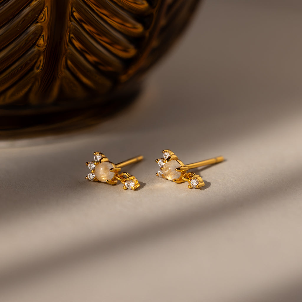 The Halo Moonstone Drop Studs in 18K Gold exude royal elegance with moonstone and crystal accents, displayed on a light surface beside a decorative golden object.