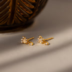 The Halo Moonstone Drop Studs in 18K Gold exude royal elegance with moonstone and crystal accents, displayed on a light surface beside a decorative golden object.