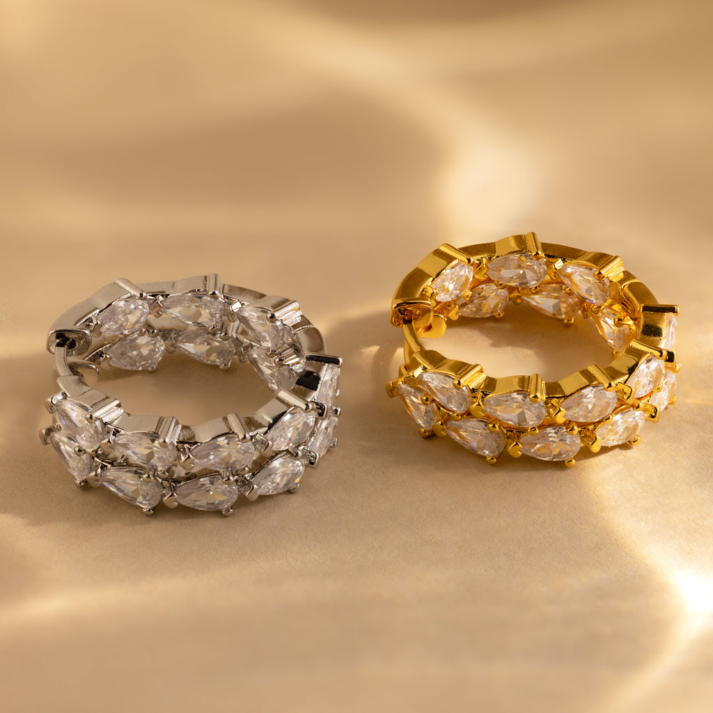 Two gemstone-encrusted rings, one silver and one gold, sit on a soft beige surface with warm lighting, sparkling beside the elegant Inside Out Pear Diamond Hoops for a truly sophisticated collection.