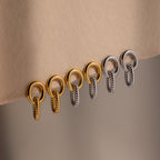 Six metallic rings, three gold and three silver, hang in a row from a beige fabric edge—mirroring the sculptural elegance of the Twisted Link Earrings.