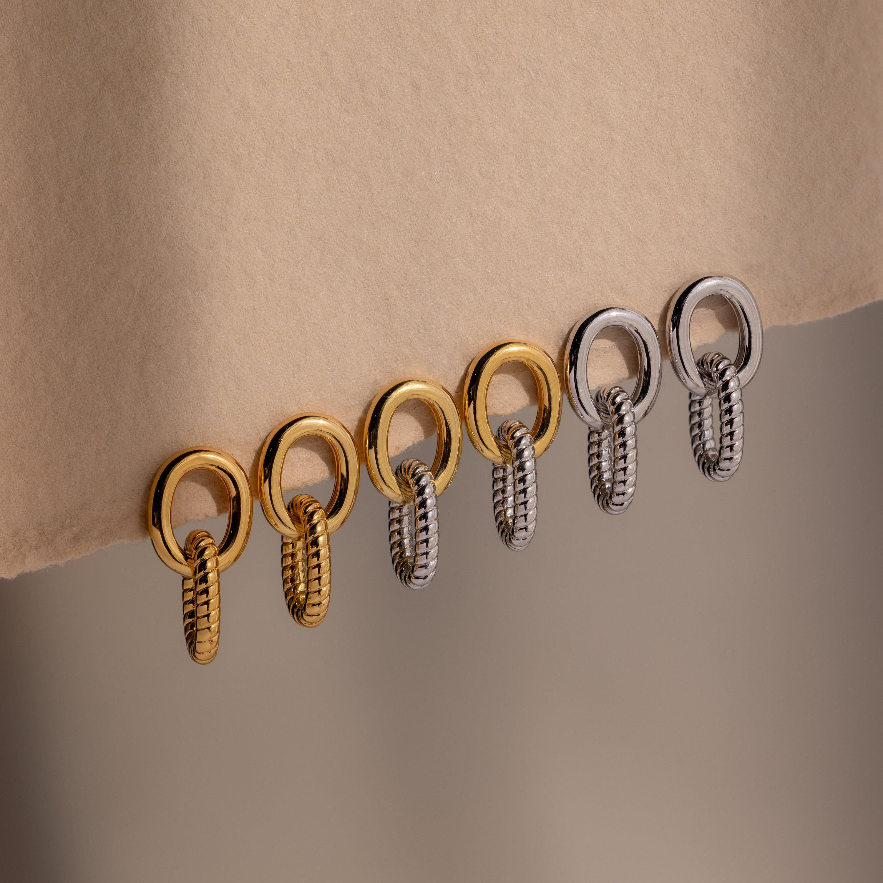 Six metallic rings, three gold and three silver, hang in a row from a beige fabric edge—mirroring the sculptural elegance of the Twisted Link Earrings.