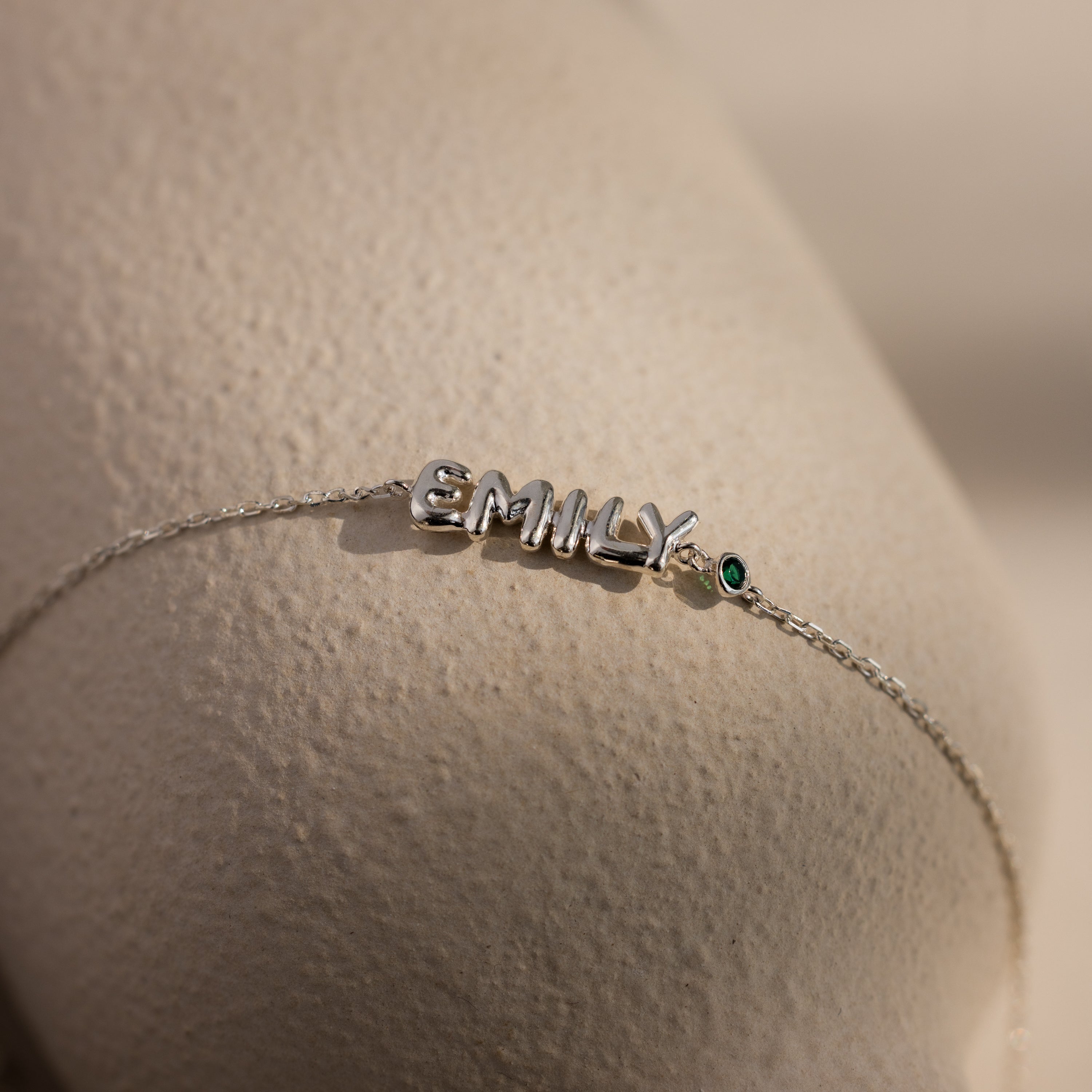 Up close of a dainty silver bubble letter bracelet with the name "EMILY" and a green emerald birthstone, displayed against a neutral background.