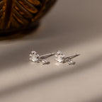 A pair of Halo Moonstone Drop Studs, featuring silver with small pearls and sparkling stones, are displayed on a light surface beside a patterned brown item.