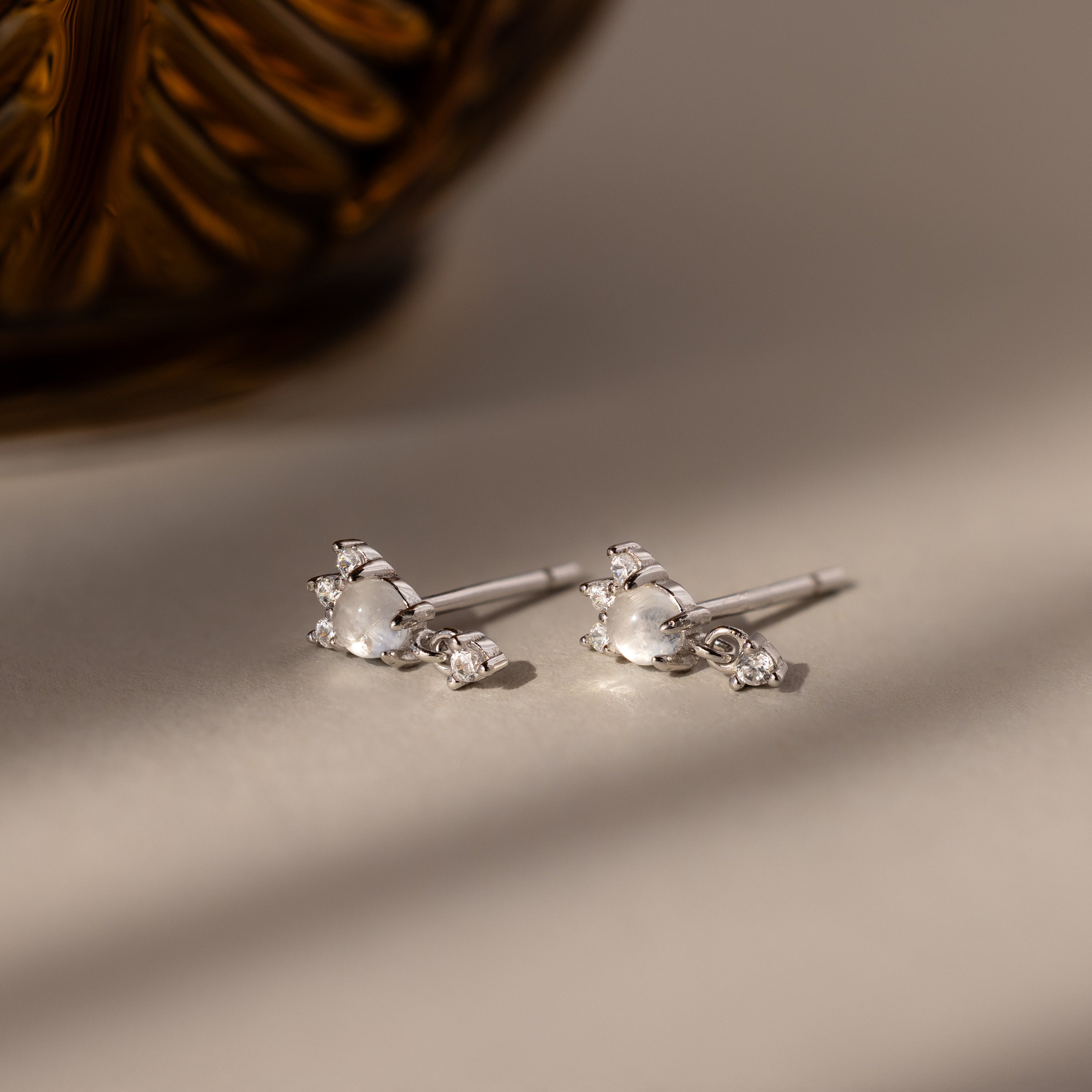 A pair of Halo Moonstone Drop Studs, featuring silver with small pearls and sparkling stones, are displayed on a light surface beside a patterned brown item.