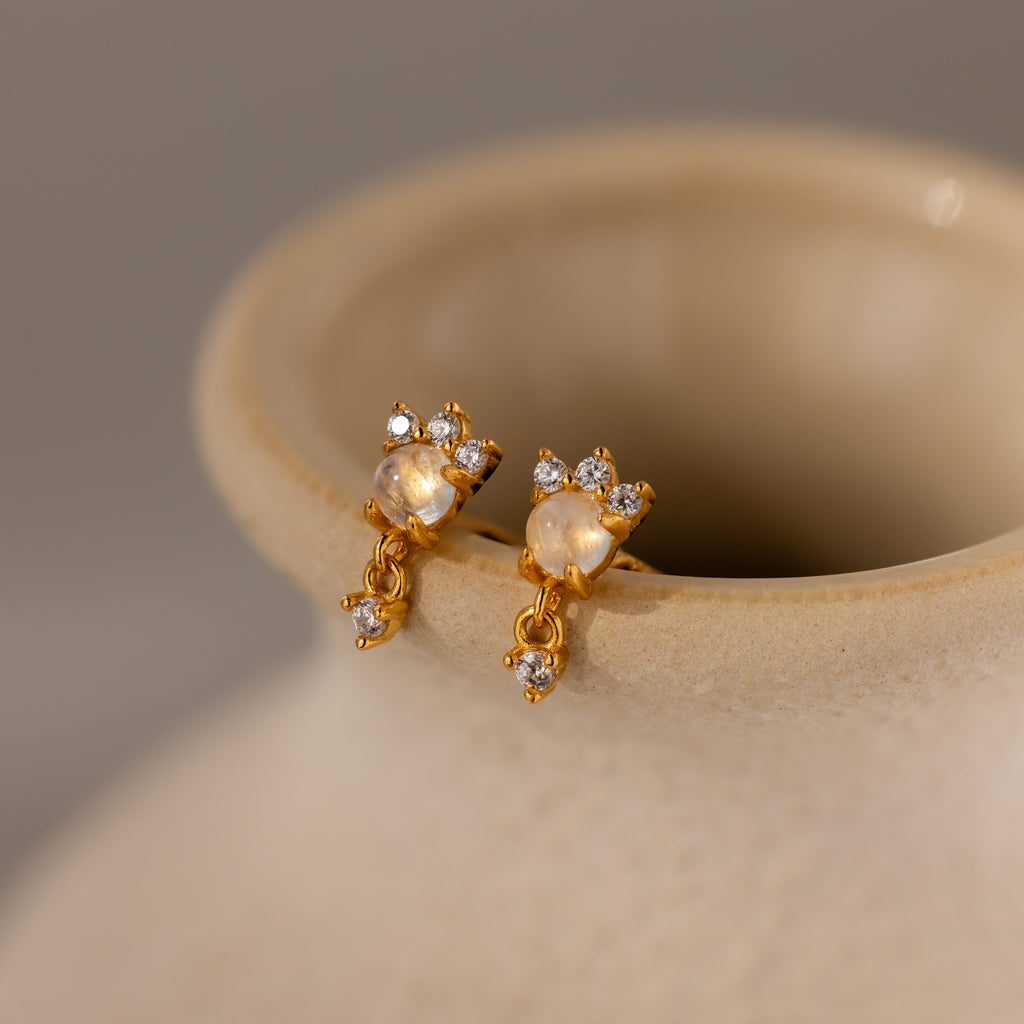 The Halo Moonstone Drop Studs in 18K Gold exude royal elegance, just like small crystals glimmering on the rim of a beige ceramic vase.