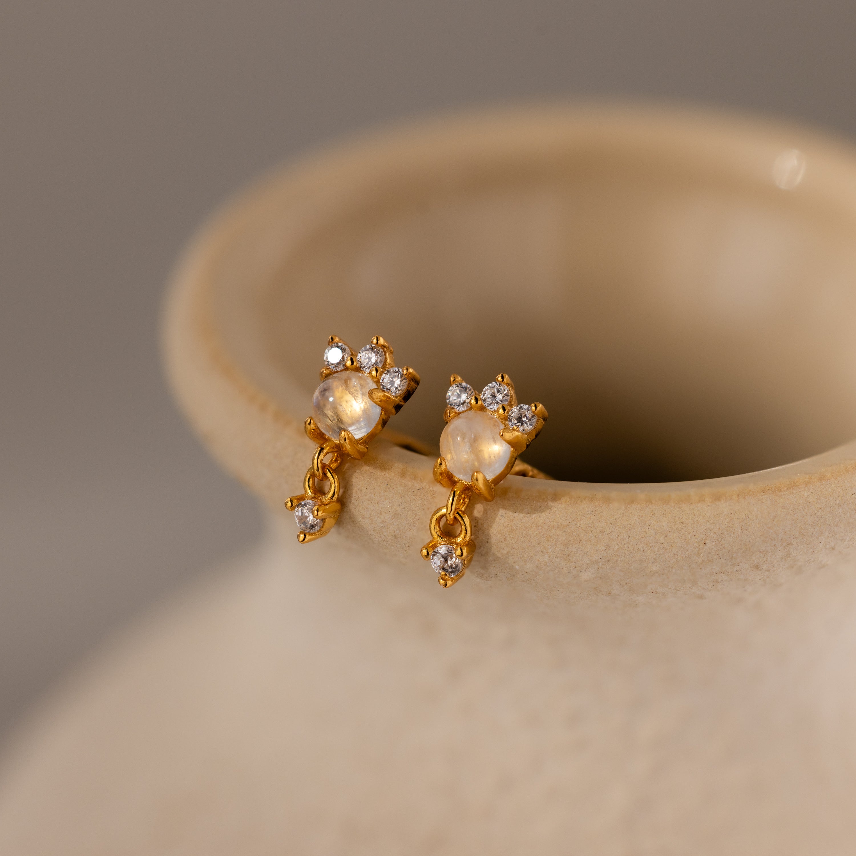 The Halo Moonstone Drop Studs in 18K Gold exude royal elegance, just like small crystals glimmering on the rim of a beige ceramic vase.