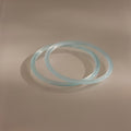 Two thin blue jade bangles from the Thin Blue Jade Bangle Set overlap on a smooth, neutral surface under soft lighting, highlighting their delicate design.