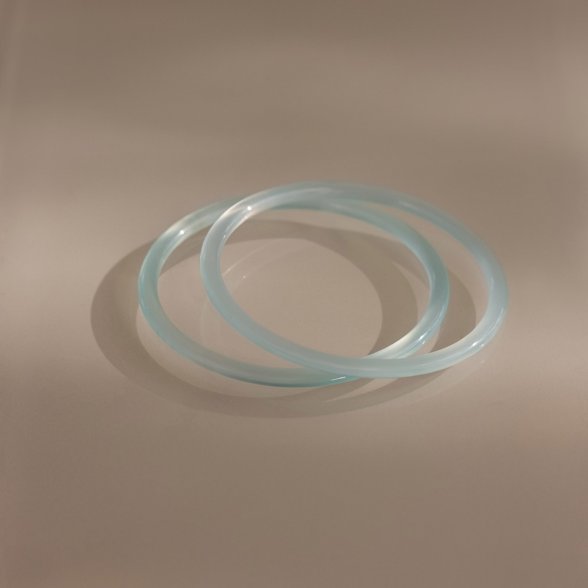 Two thin blue jade bangles from the Thin Blue Jade Bangle Set overlap on a smooth, neutral surface under soft lighting, highlighting their delicate design.