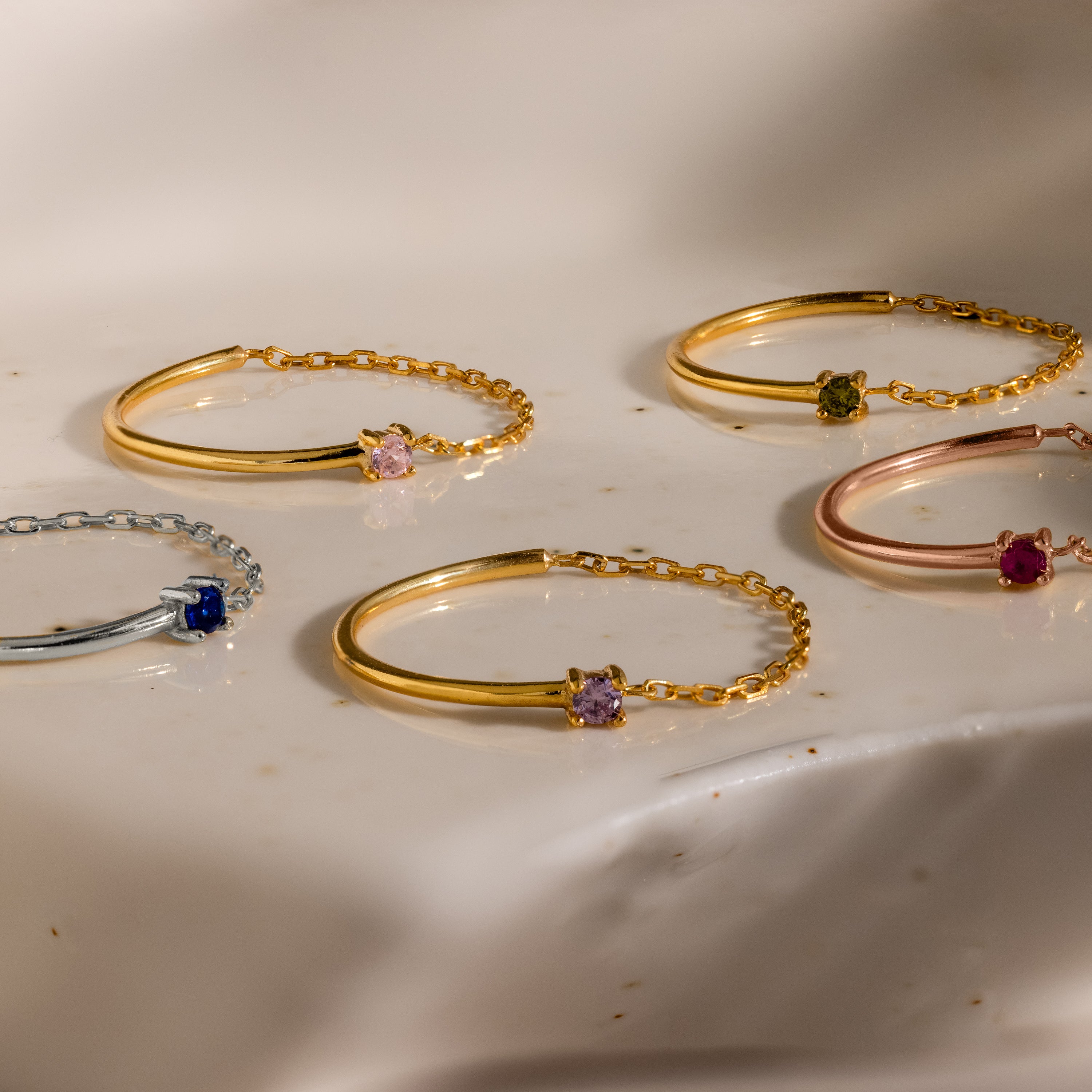 Five delicate chain bracelets in gold, silver, and rose gold with colorful gemstones rest on marble—perfect to pair with the Birthstone Half Chain Ring or other dainty jewelry for a subtle, elegant look.