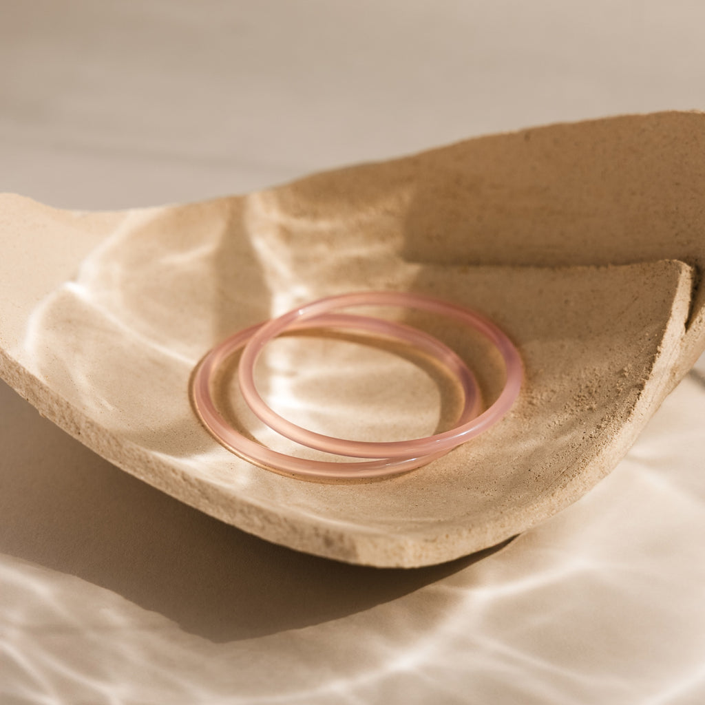 Two bangles from the Thin Pink Jade Bangle Set rest on a textured beige ceramic dish, bathed in soft natural light—ideal for layering and adding elegance to your bracelet stack.