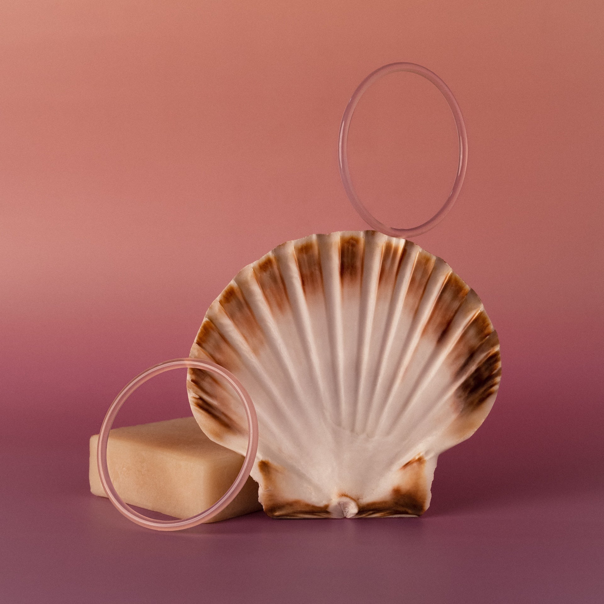 The Thin Pink Jade Bangle Set is artfully arranged with a seashell and bar of soap on a pink gradient background.