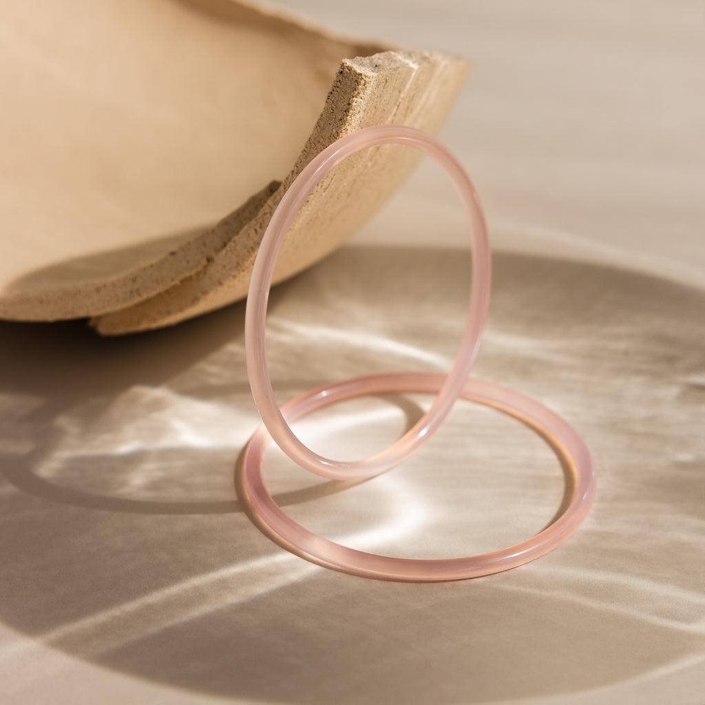 The Thin Pink Jade Bangle Set features two translucent pink jade bangles displayed on a light surface beside a beige ceramic bowl—ideal for effortless layering.