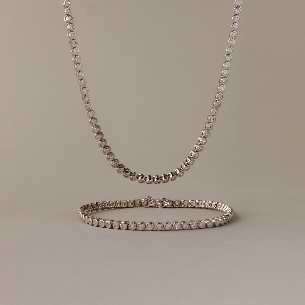 The Diamond Bezel Tennis Necklace & Bracelet Set in White Gold, displayed on a beige background, makes an elegant and unforgettable anniversary gift.