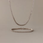 The Diamond Bezel Tennis Necklace & Bracelet Set in White Gold, displayed on a beige background, makes an elegant and unforgettable anniversary gift.