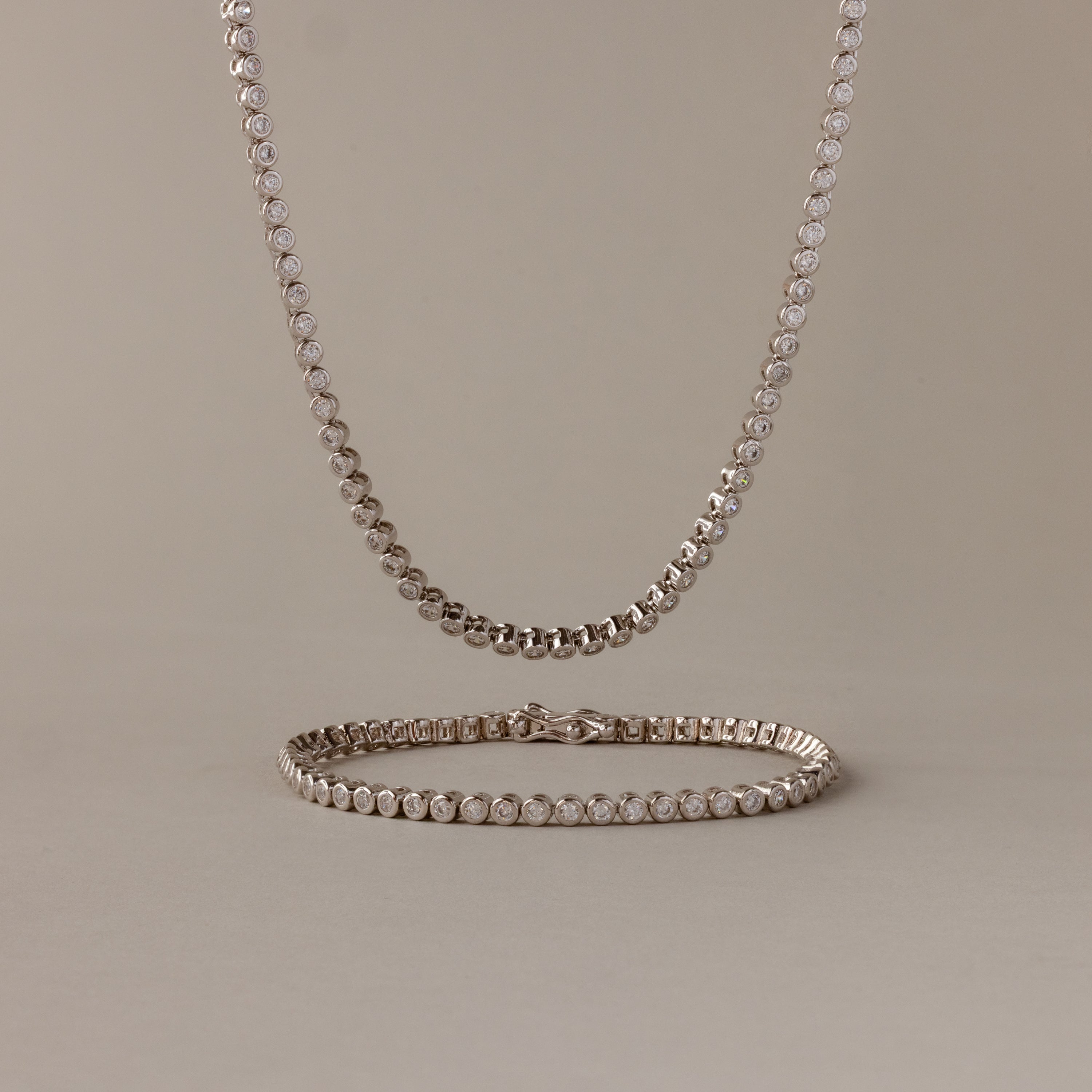 The Diamond Bezel Tennis Necklace & Bracelet Set in White Gold, displayed on a beige background, makes an elegant and unforgettable anniversary gift.