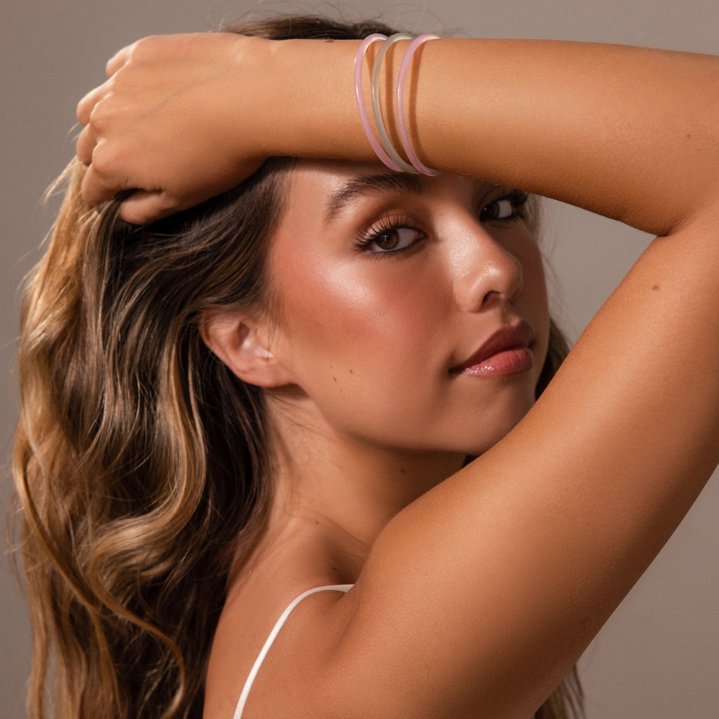 Woman with wavy hair poses with arm raised, wearing a white strap top and showcasing the Thin Pink Jade Bangle Set, looking at the camera.