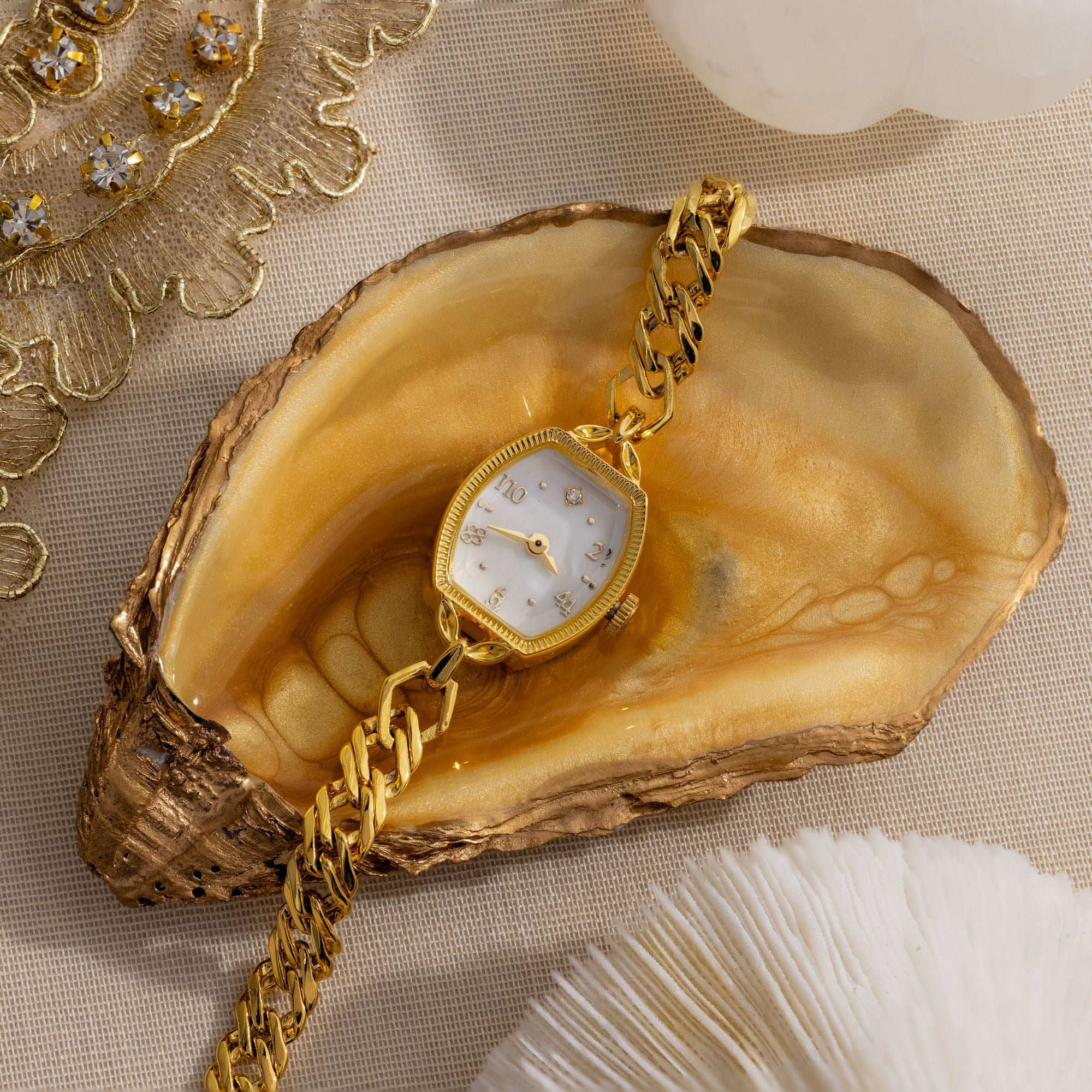 The Heirloom Chain Link Timepiece, featuring a mother of pearl dial, is showcased on an open oyster shell with a gold-trimmed edge.