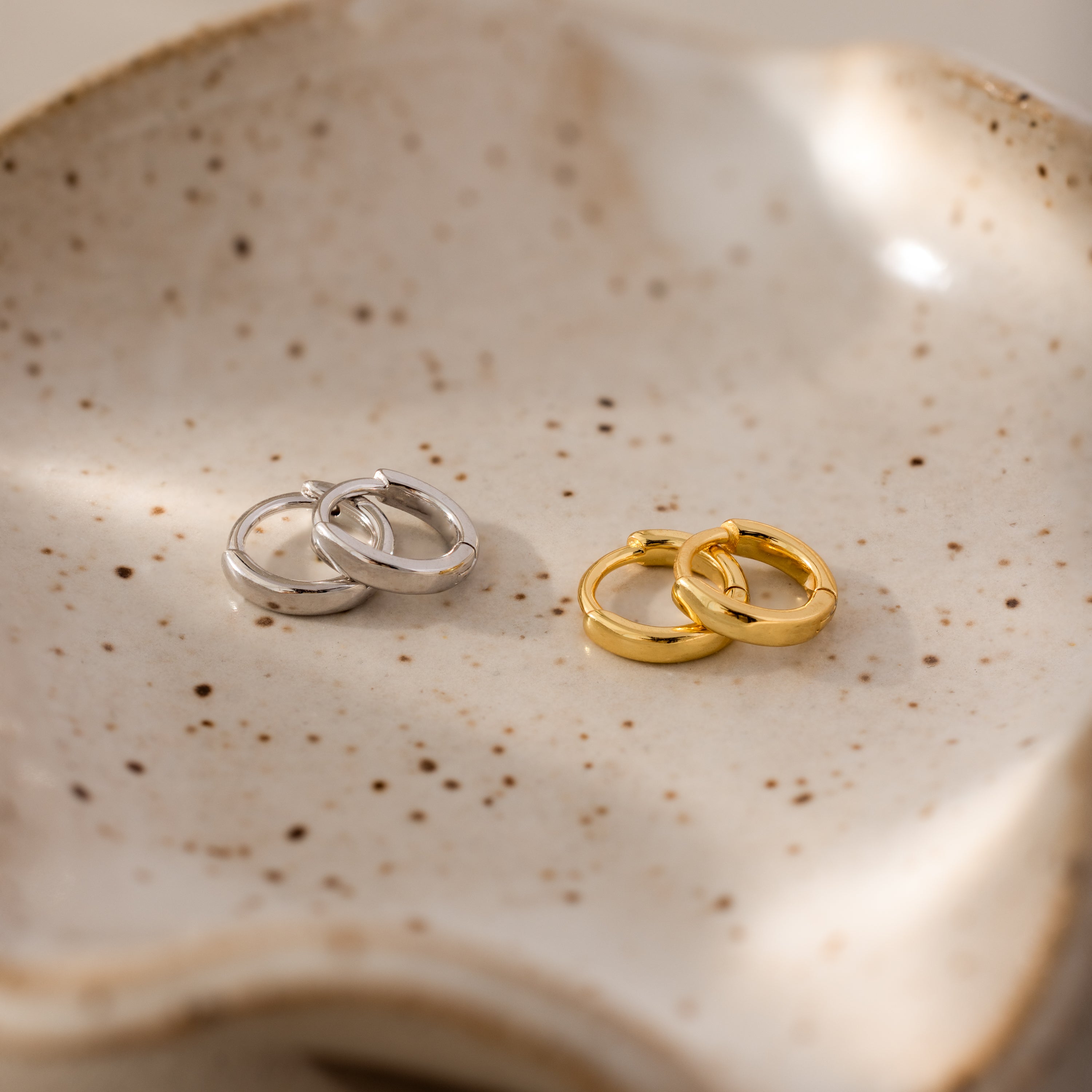 Pair of 6mm tiny huggie hoop earrings in gold and silver finishes, displayed on a speckled ceramic dish.