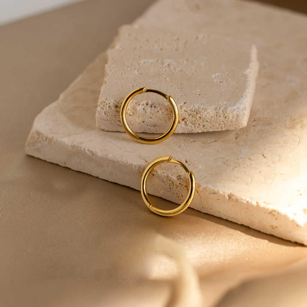 The Medium Bold Hoops are stylish gold earrings, lightweight and perfect for everyday wear, shown on textured beige stone slabs with a soft neutral background.