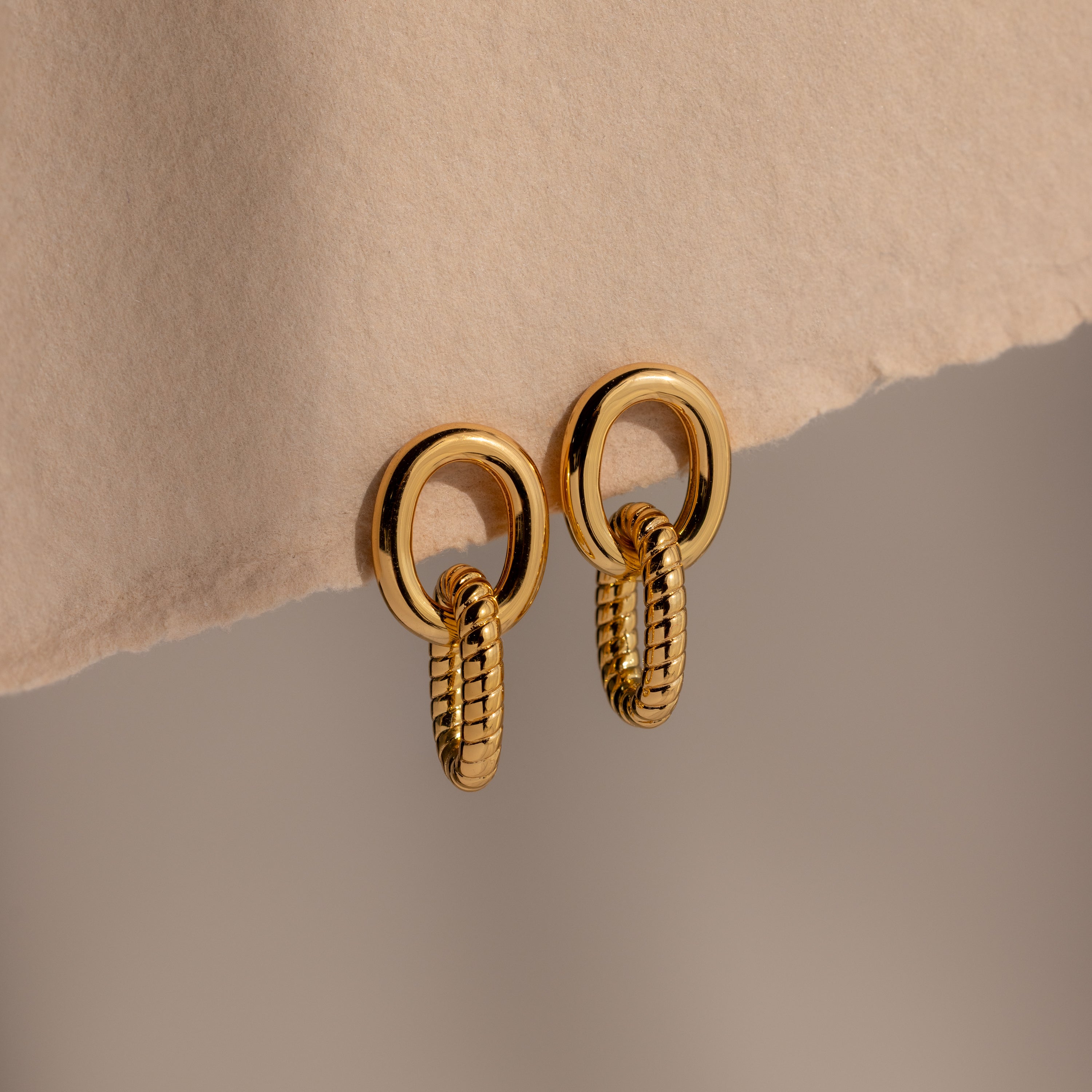 Twisted Link Earrings in 18K Gold showcase an oval, textured chain design against a soft beige background—ideal for those who love modern and elegant jewelry.