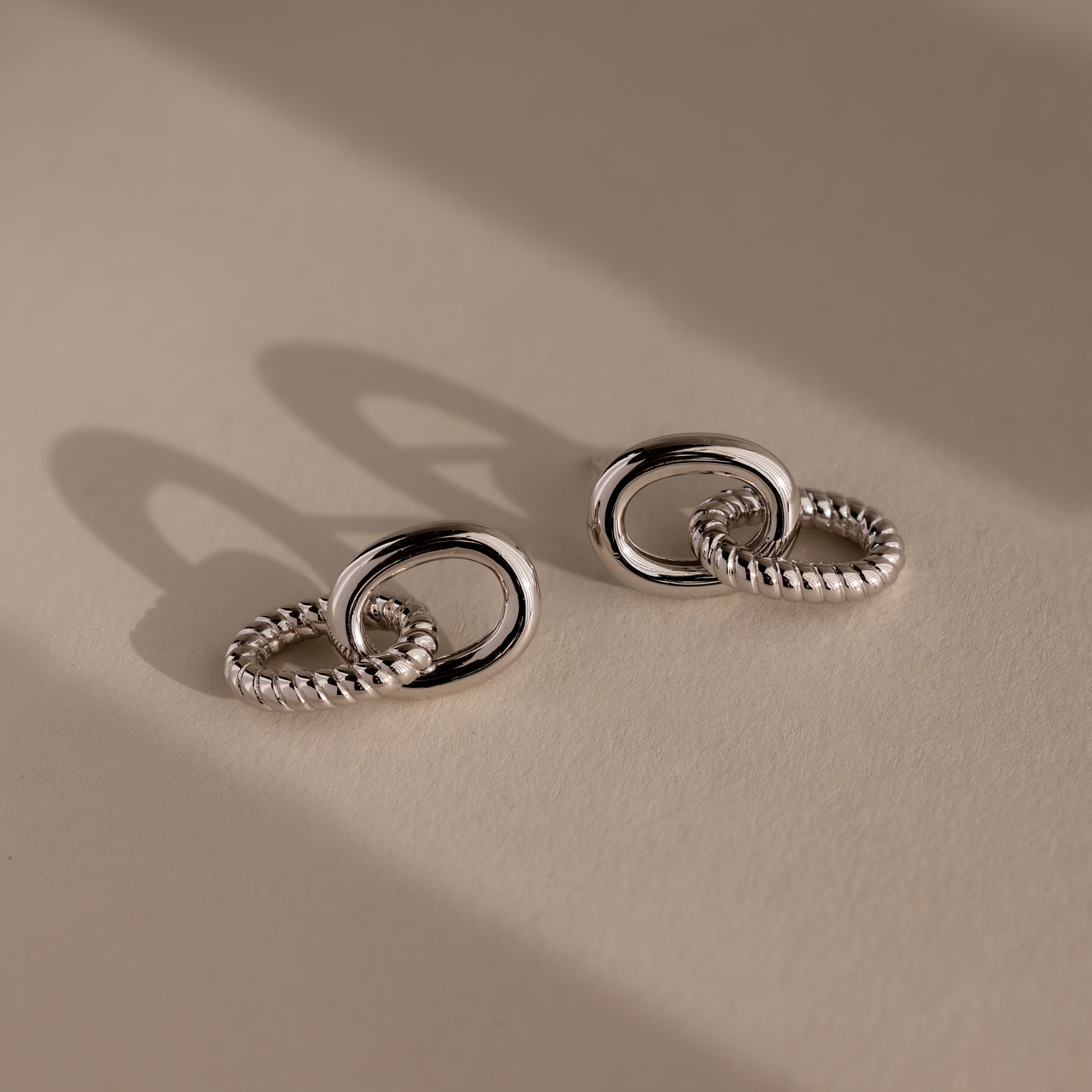 Two pairs of Twisted Link Earrings in White Gold, these modern earrings feature intertwined hoops casting soft shadows on a neutral background—a perfect example of timeless jewelry.