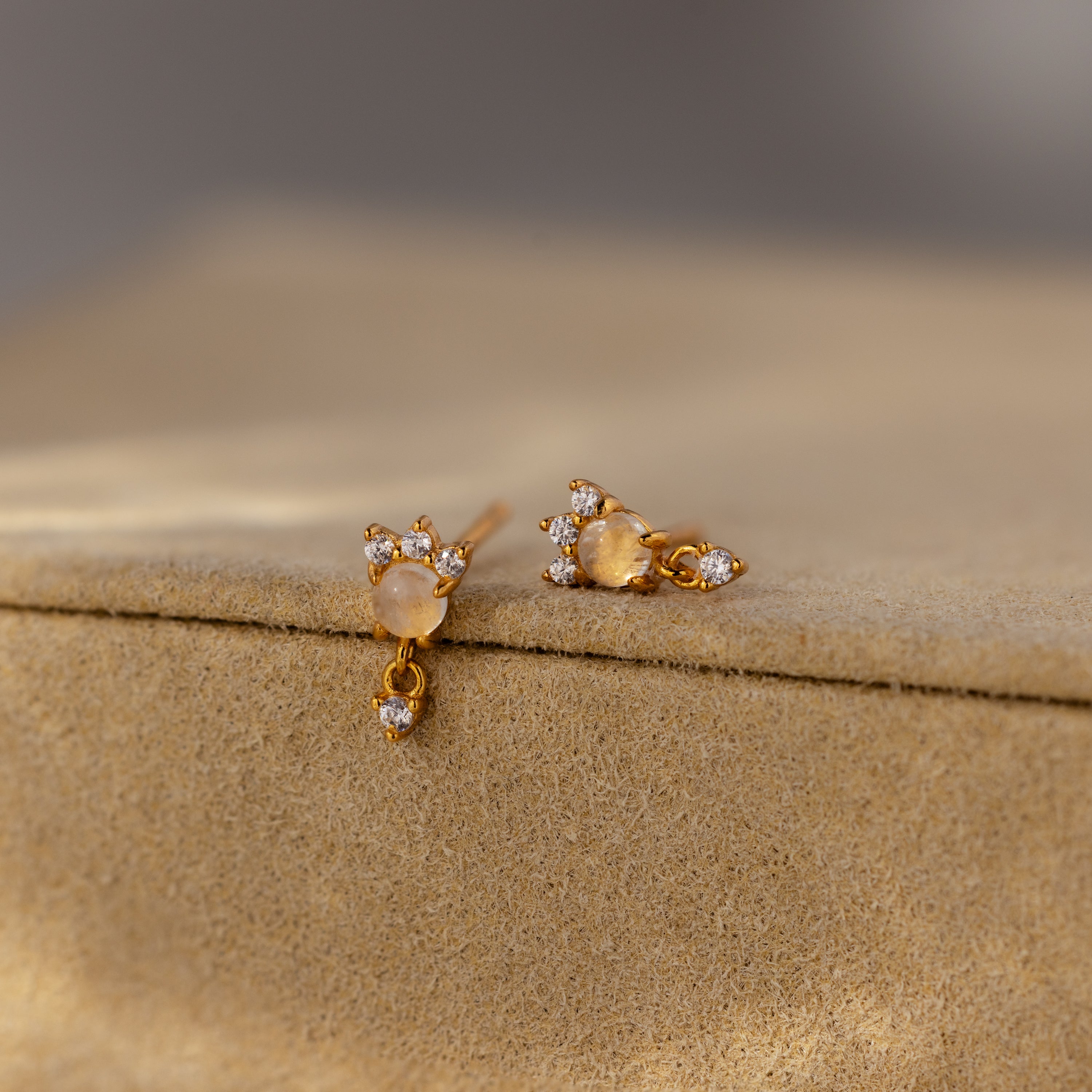 Halo Moonstone Drop Studs featuring small white gemstones and a dangling accent, displayed on a beige textured surface.