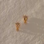 The Halo Moonstone Drop Studs in 18K Gold radiate royal elegance, their round stones shimmering against a textured cream backdrop.