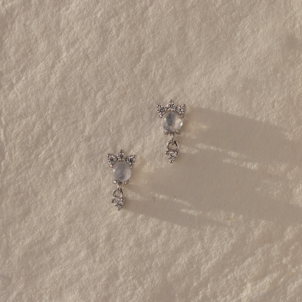 The Halo Moonstone Drop Studs in Sterling Silver exude royal elegance, their star-inspired design shimmering beautifully on a textured, light surface.