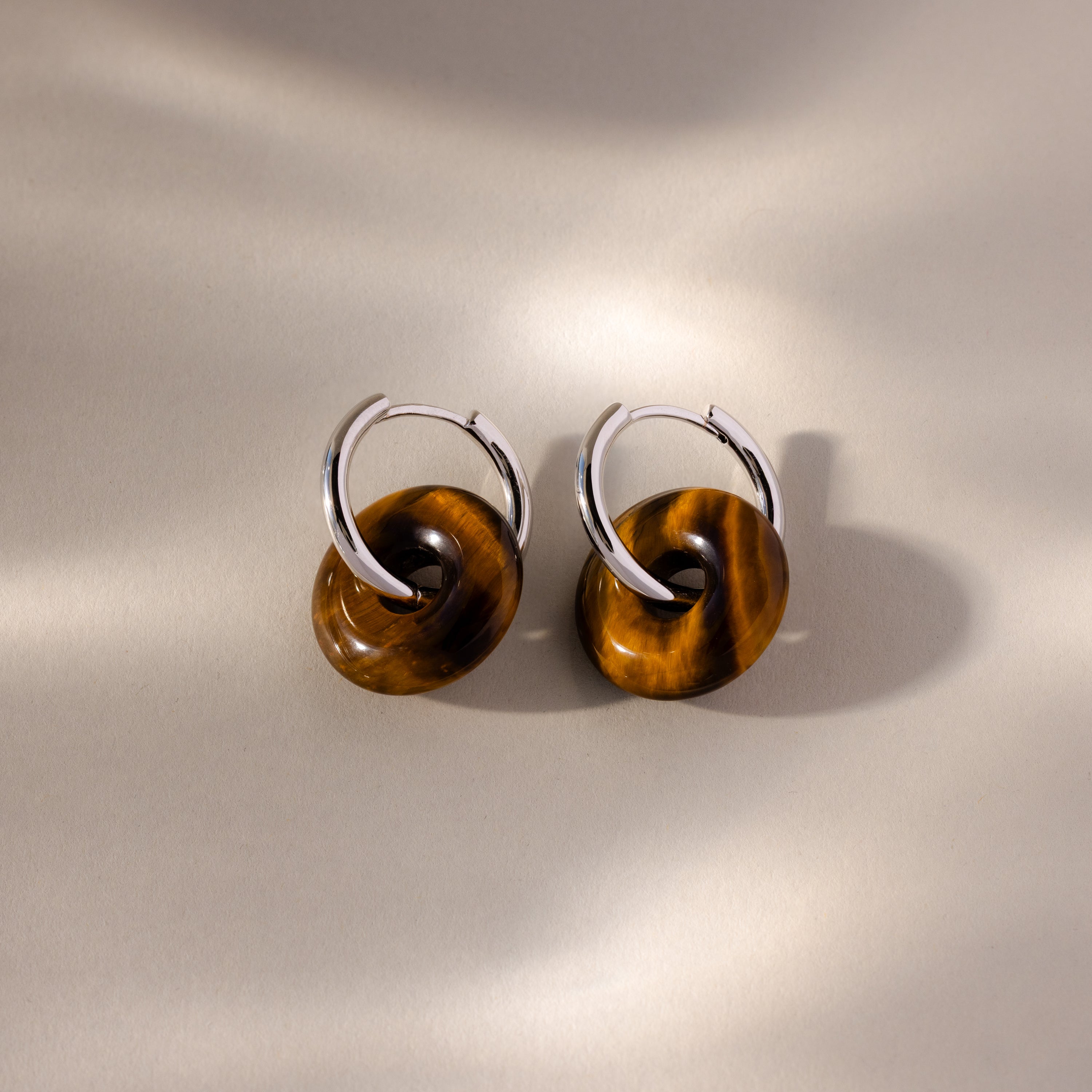 Tiger's Eye Duo Hoops in White Gold, featuring brown circular stone charms, are displayed on a neutral background with soft shadows.