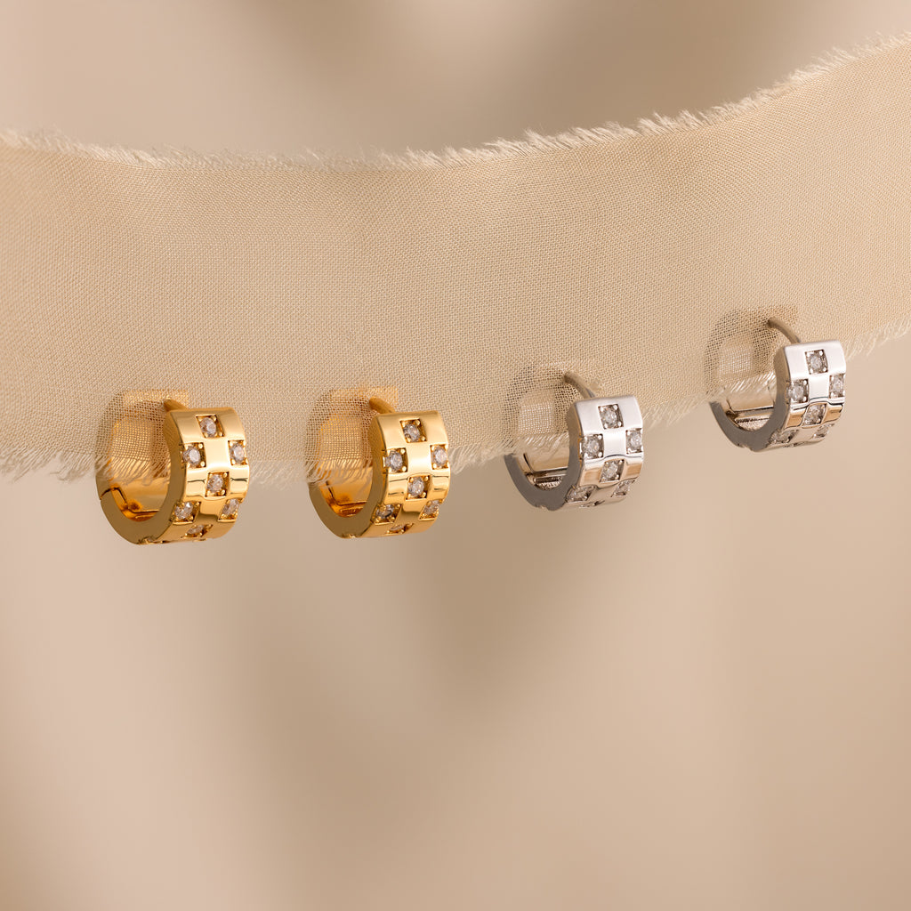 Four hoop earrings, featuring chunky hoops and Pave Checkered Huggies, in gold and silver with square and round details, are shown on beige fabric.