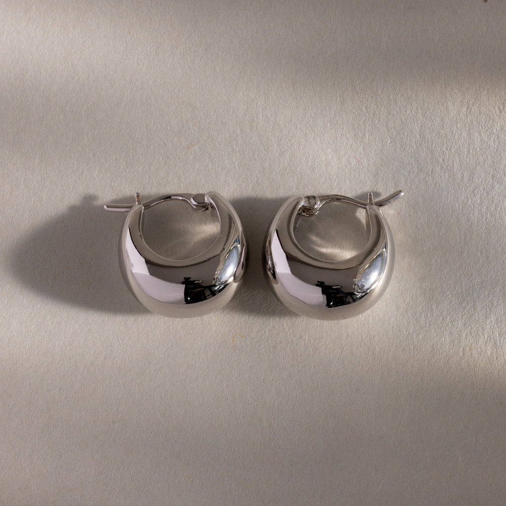 The Bold Teardrop Earrings in White Gold showcase a shiny teardrop silhouette, displayed on a light textured surface with soft shadows.