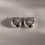 The Bold Teardrop Earrings in White Gold showcase a shiny teardrop silhouette, displayed on a light textured surface with soft shadows.