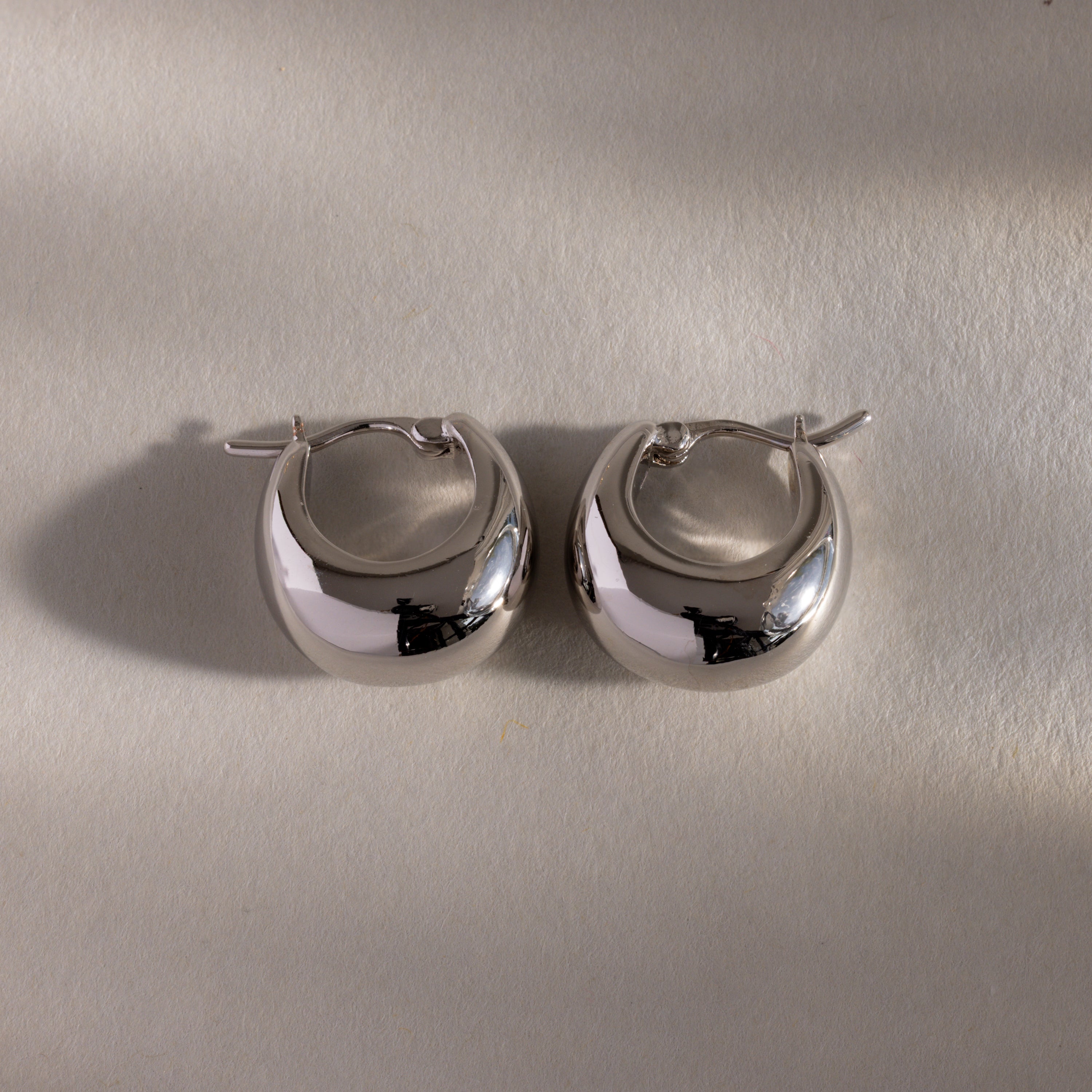 The Bold Teardrop Earrings in White Gold showcase a shiny teardrop silhouette, displayed on a light textured surface with soft shadows.