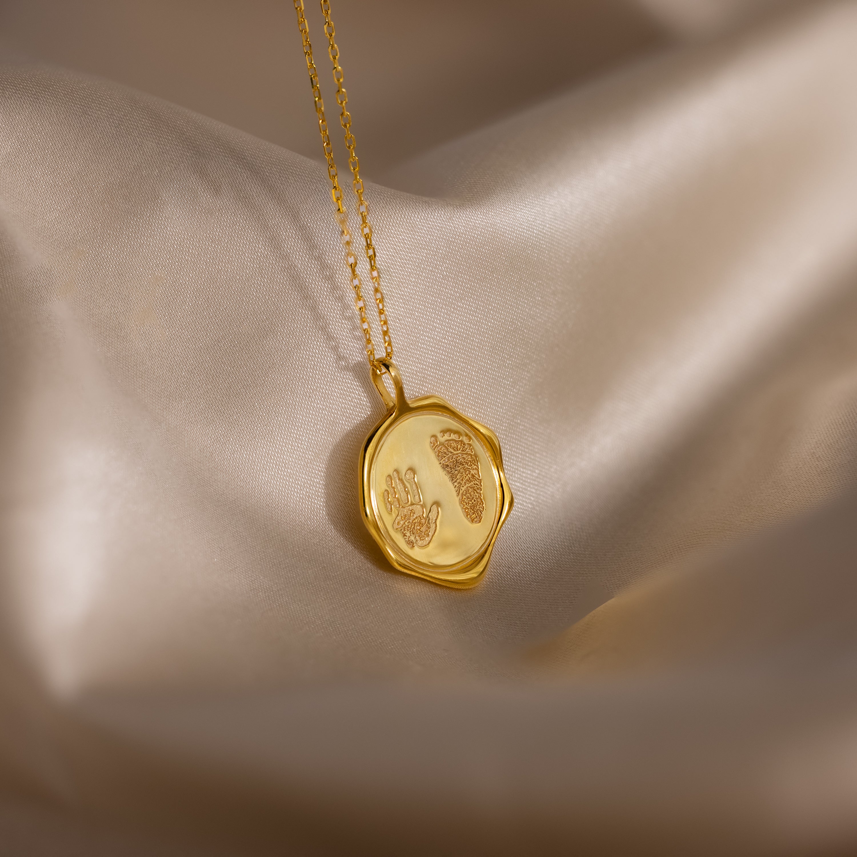 The Baby Hand & Footprint Necklace in 18K Gold features an intricate baby hand and footprint design, elegantly presented on soft beige satin fabric.