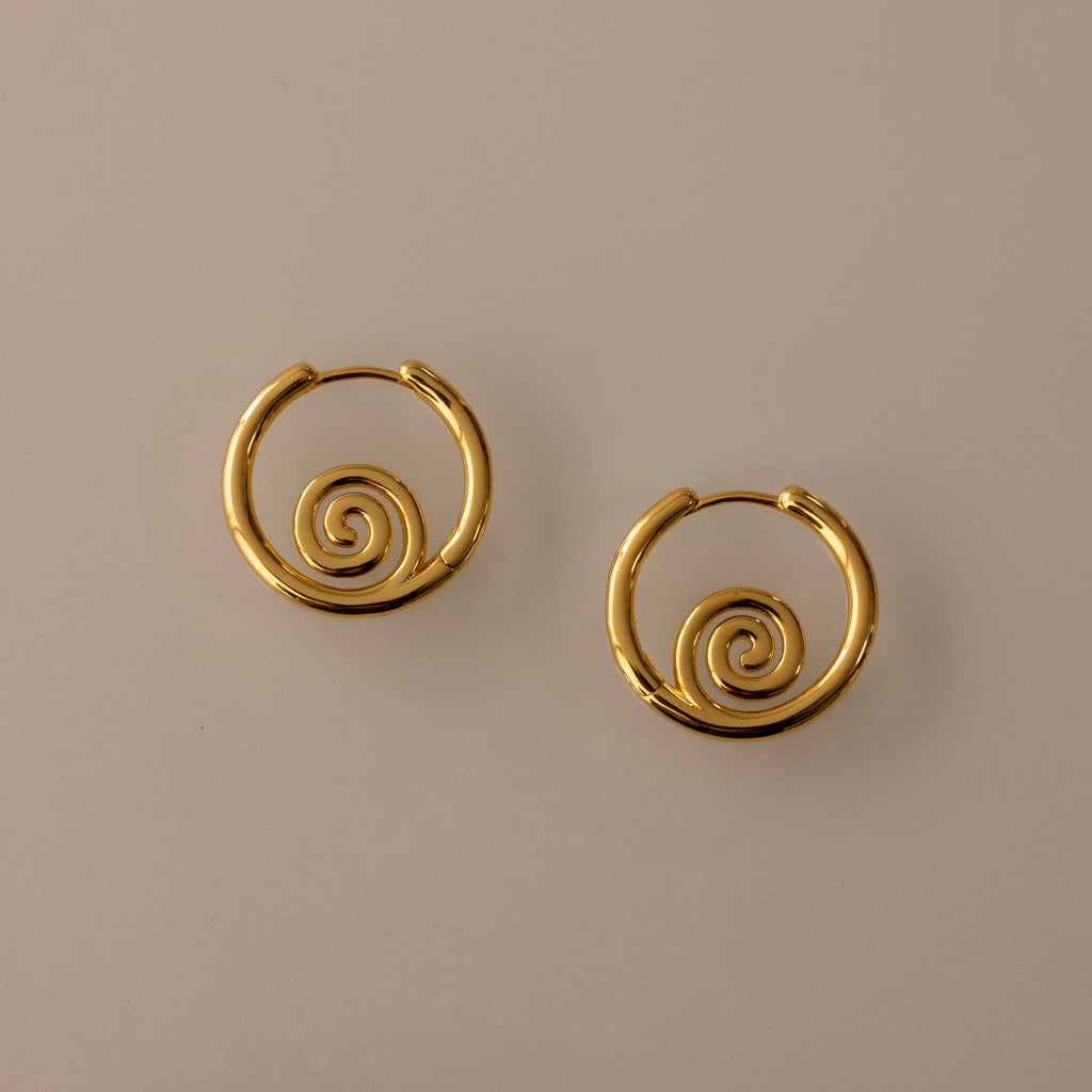 The Spiral Swirl Hoops in 18K Gold feature intricate spiral designs, making them perfect boho vacation jewelry, shown against a plain beige background.