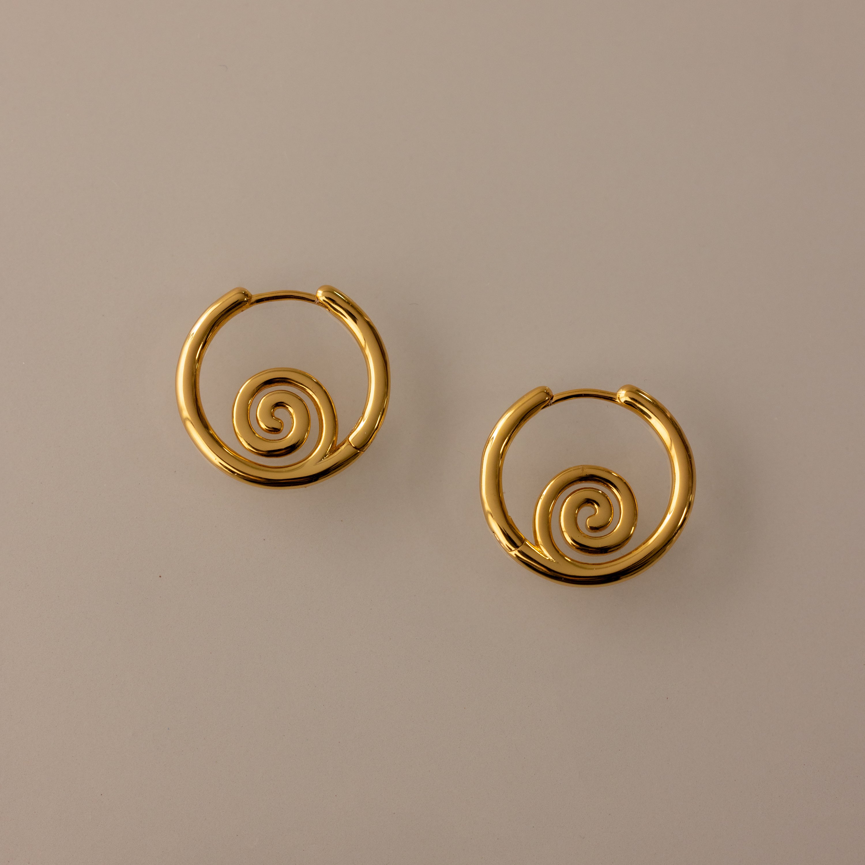 The Spiral Swirl Hoops in 18K Gold feature intricate spiral designs, making them perfect boho vacation jewelry, shown against a plain beige background.