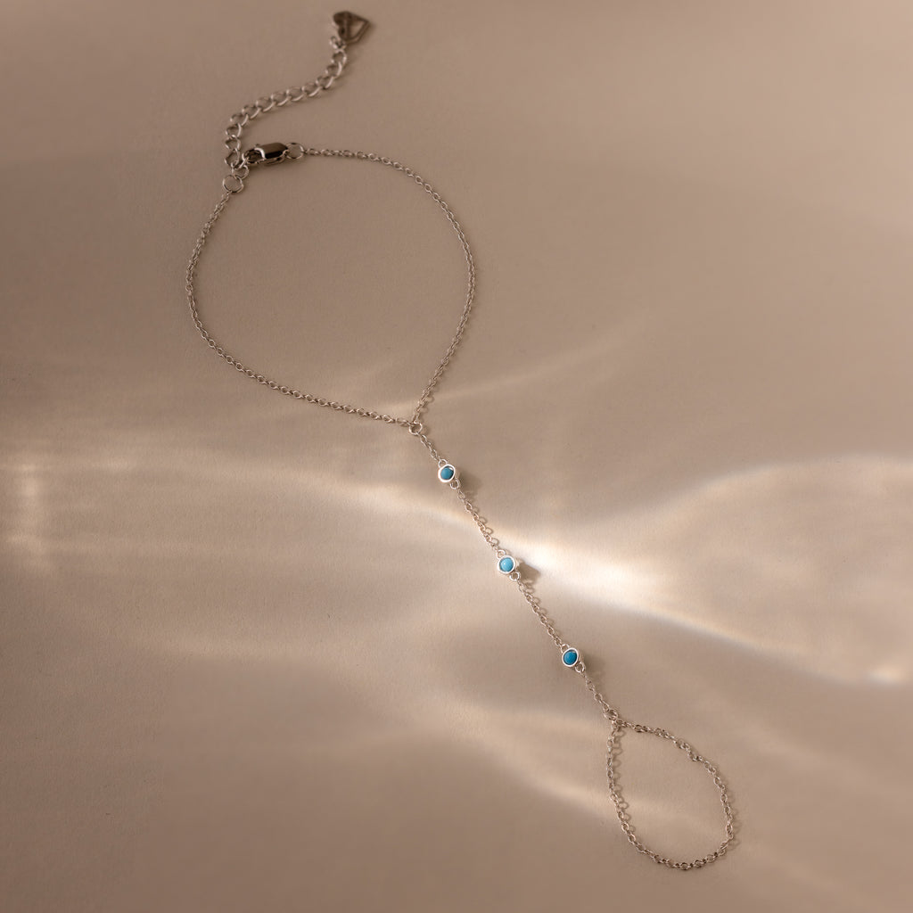 The Turquoise Hand Chain Bracelet in Silver is shown on a beige surface, featuring delicate silver links and turquoise stones highlighted by soft reflected light.