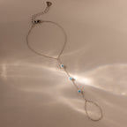 The Turquoise Hand Chain Bracelet in Silver is shown on a beige surface, featuring delicate silver links and turquoise stones highlighted by soft reflected light.