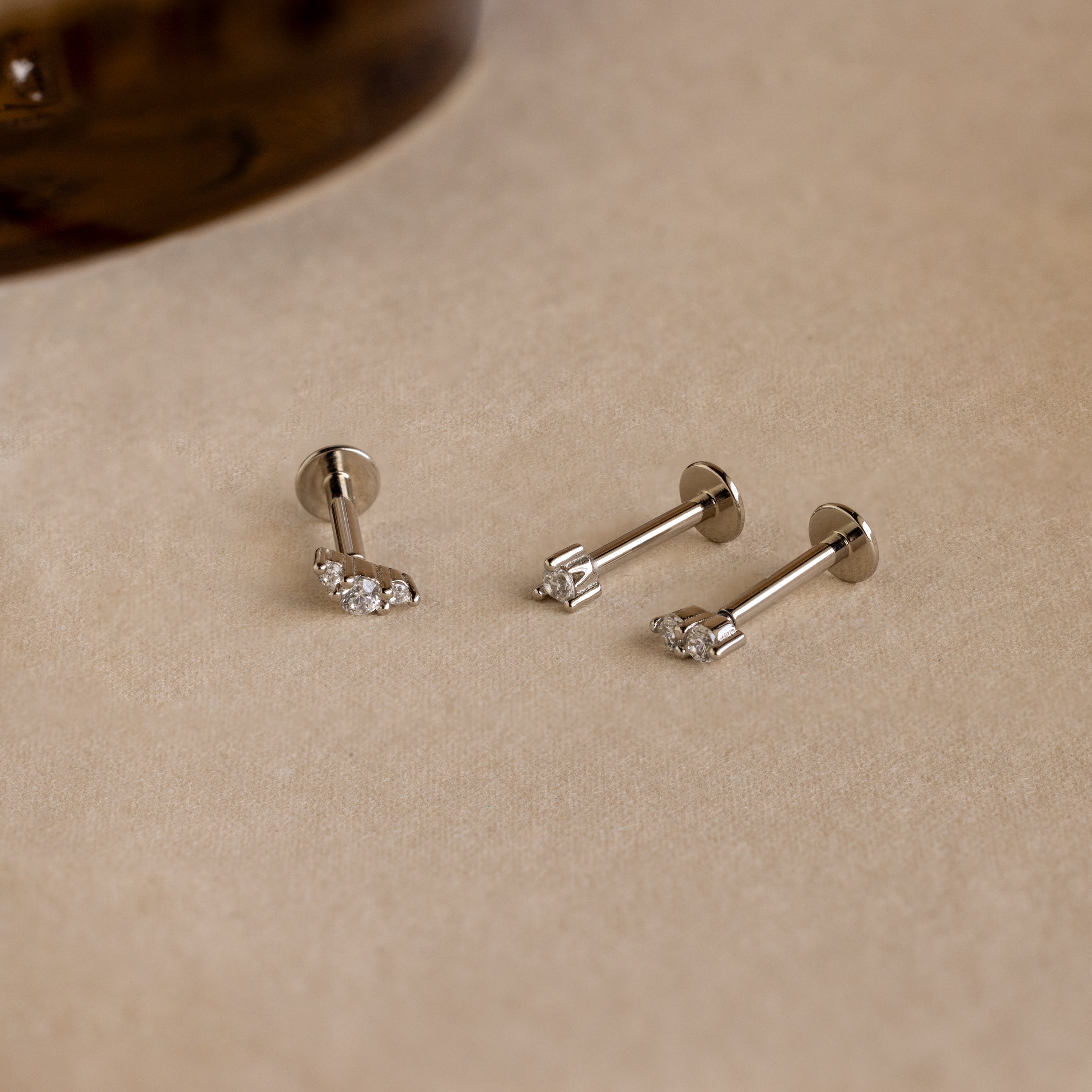 Three silver Tiny Diamond Flatback Studs from the set, featuring clear gemstones, are displayed on a light beige surface—ideal for those who love classic diamond stud earrings.