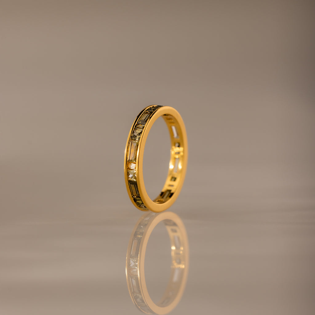 The Peridot Inlay Eternity Ring with gold and decorative patterns stands upright, its gleaming reflection visible on a smooth, neutral surface.