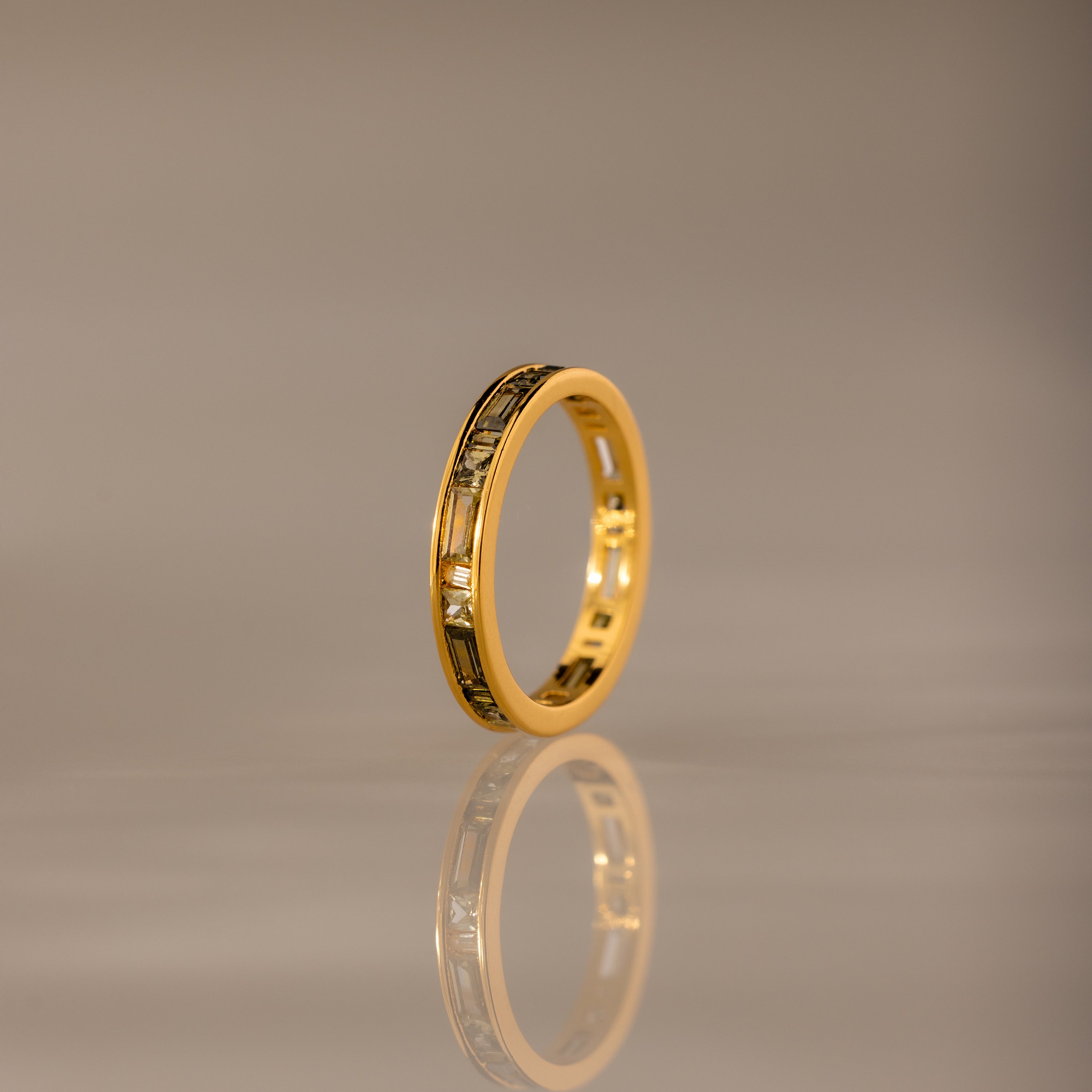 The Peridot Inlay Eternity Ring with gold and decorative patterns stands upright, its gleaming reflection visible on a smooth, neutral surface.